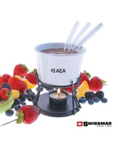 A white ceramic fondue bowl filled with chocolate and the forks are inside. The bowl is on a metal stand with a light below and surrounded by fruit