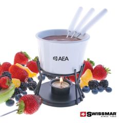 A white ceramic fondue bowl filled with chocolate and the forks are inside. The bowl is on a metal stand with a light below and surrounded by fruit