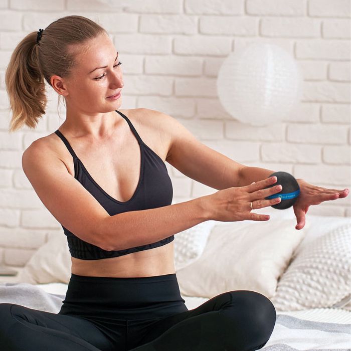 A black massage ball with a blue silicone band featuring white print. The ball is being used by a blonde woman sitting with her arms outstretched to the right in a white room.