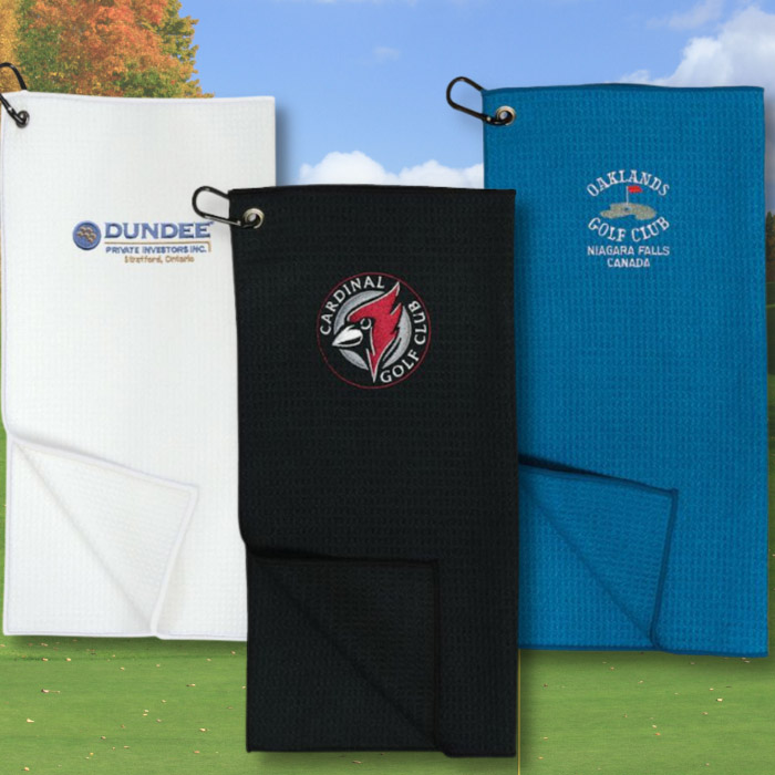 3 embroidered golf towels in white, black, and blue laid across a bright and sunny golf green.