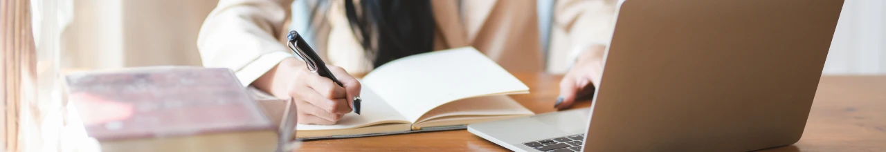 A woman with long black hair is writing in her new custom journal gift set beside her open silver-coloured laptop.